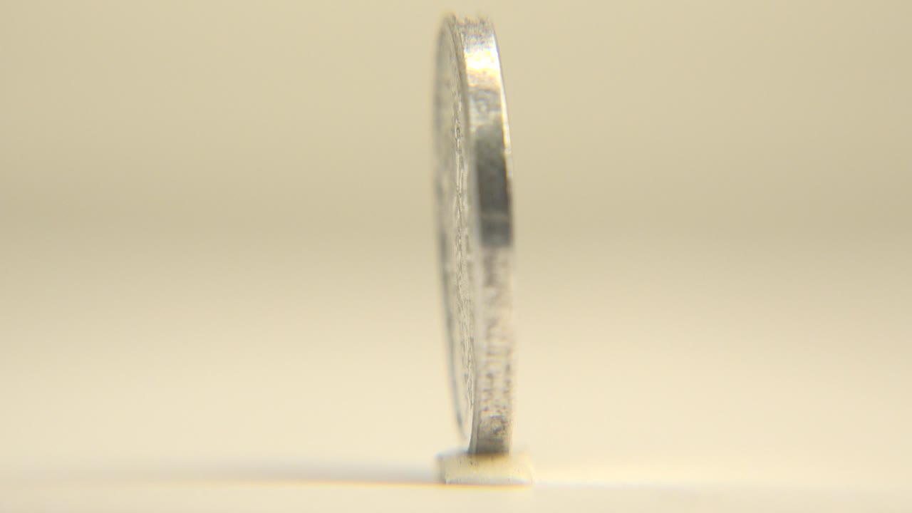 A silver coin is seen nearly edge-on at the start of the rotation, standing upright on a pale tabletop with a soft cream background and no visible people.