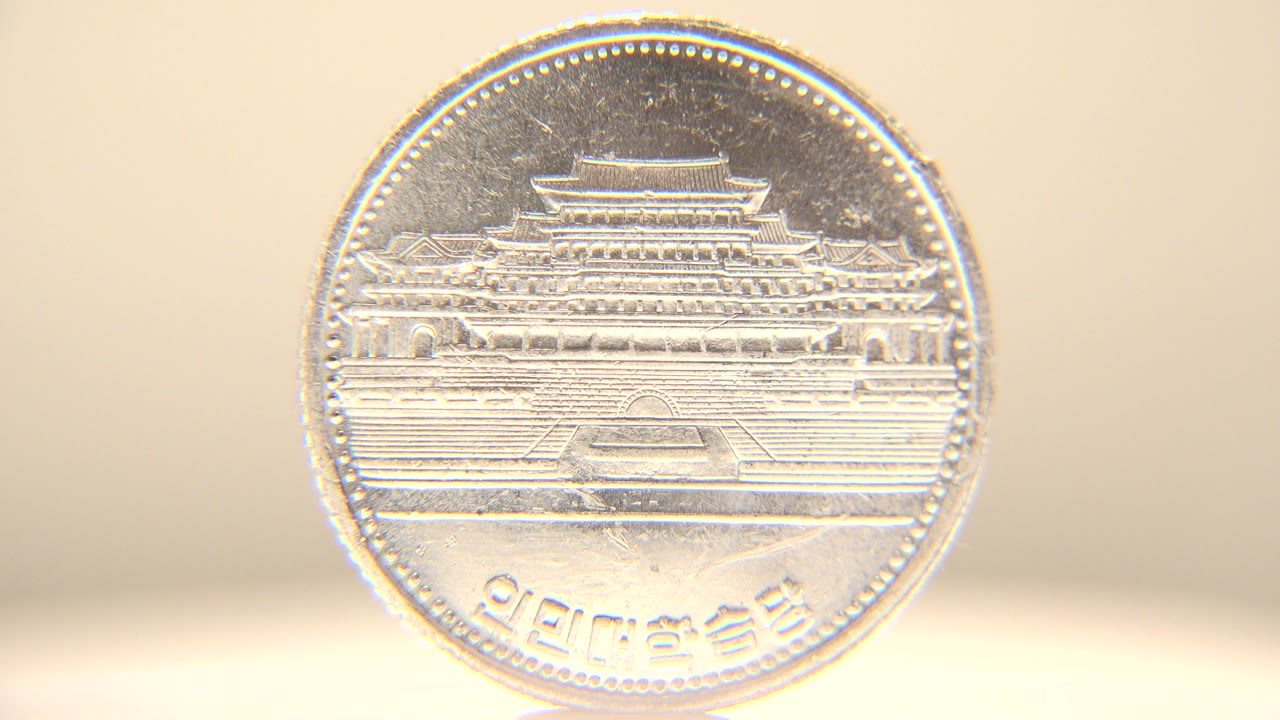 Silver coin standing upright with an embossed building design facing the camera, warm beige background, bright rim highlights, and no visible people.