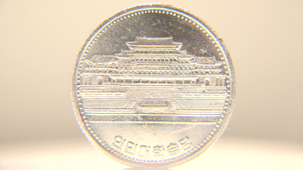 Silver coin remains nearly front-facing with the building relief and Korean characters visible, held against a soft pale background with no visible people.
