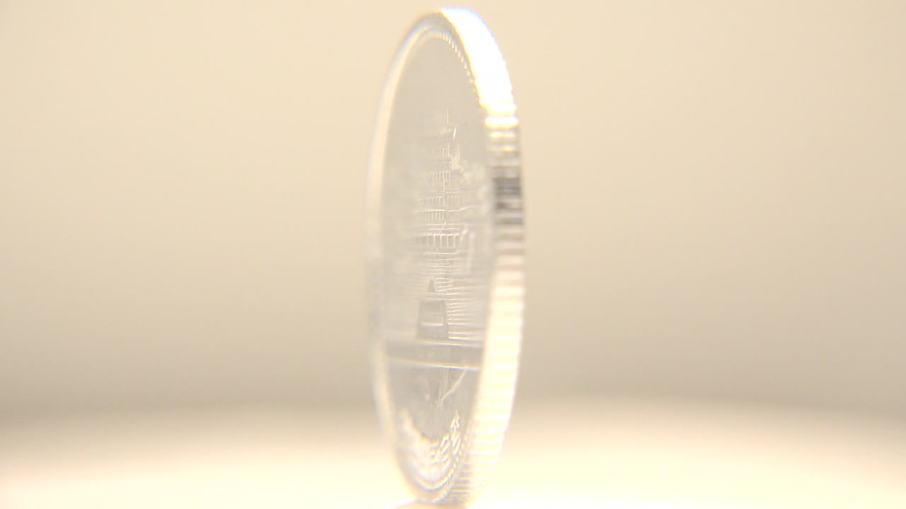 The coin rotates to a thin edge-on view, showing its ridged rim and faint reflected face against the pale tabletop, with no visible people.