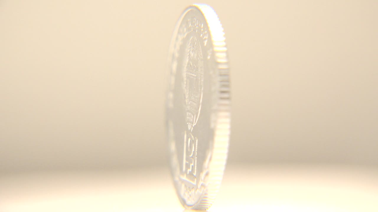 A silver-colored coin stands nearly edge-on with a bright rim highlight and soft cream background, with no visible people.