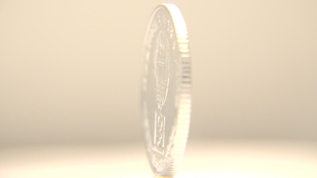 The coin remains in a narrow side view, its reflective edge catching light while the face detail stays mostly hidden, with no visible people.