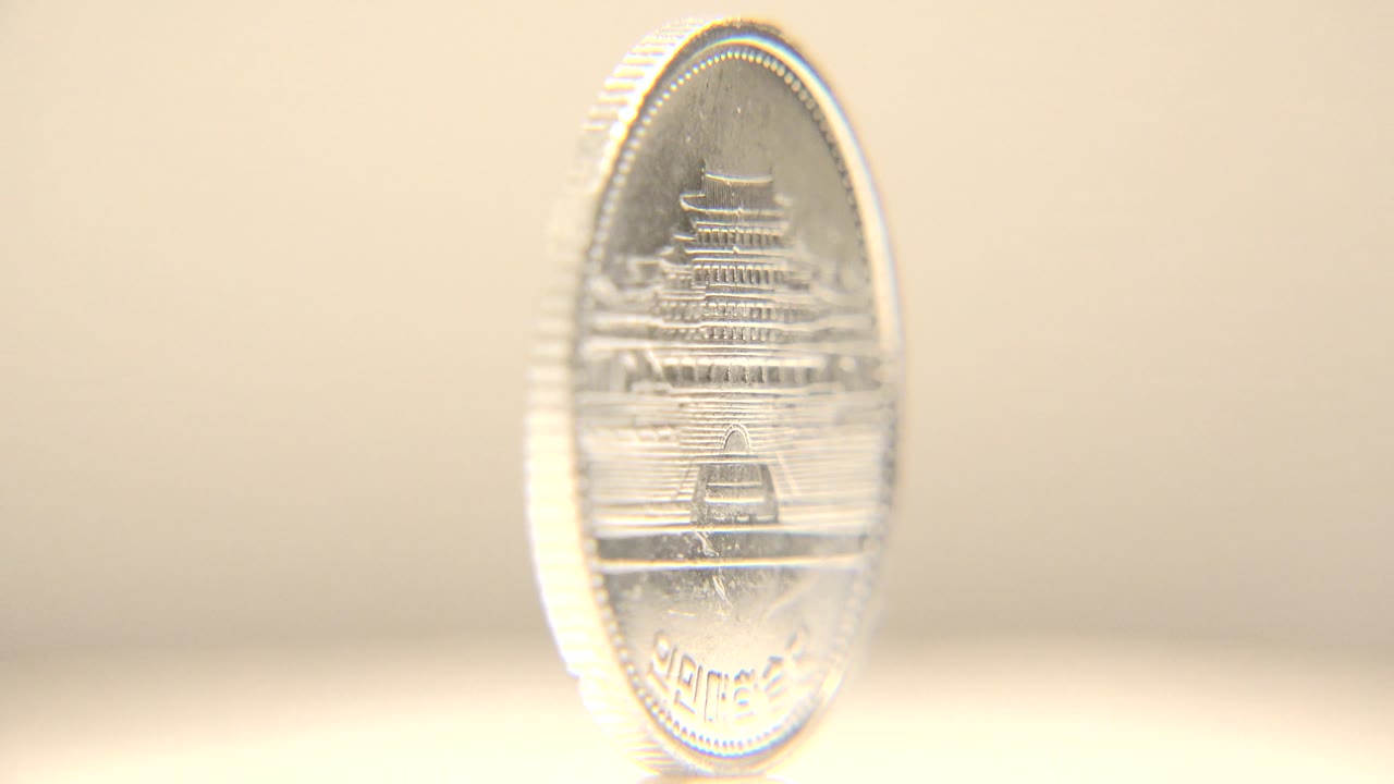 The spinning coin turns toward camera, revealing an engraved building, foreign lettering near the rim, and bright highlights on a neutral cream surface, with no visible people.
