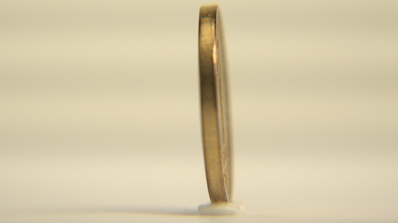 A small coin is seen nearly edge-on standing upright on a pale platform under warm light, with a soft background and no visible people.