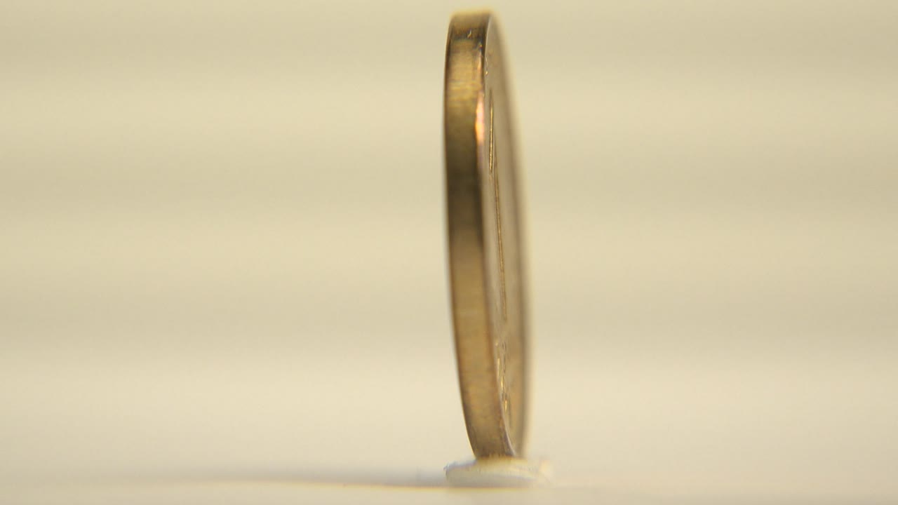 The coin remains in a thin side profile on the pale rotating surface, catching a bright vertical highlight along its rim, with no visible people.