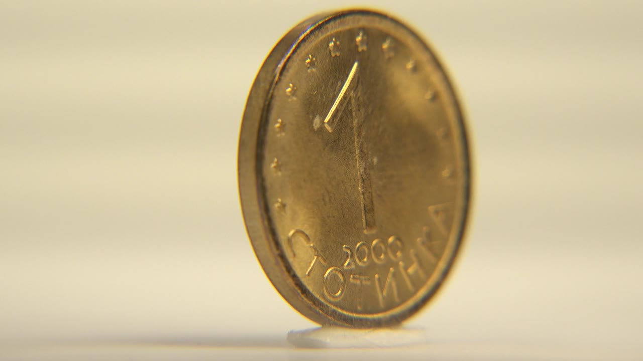 The upright coin has rotated toward a frontal angle, showing a large number one, a 2000 date, and Cyrillic lettering on a warm brass surface, with no visible people.