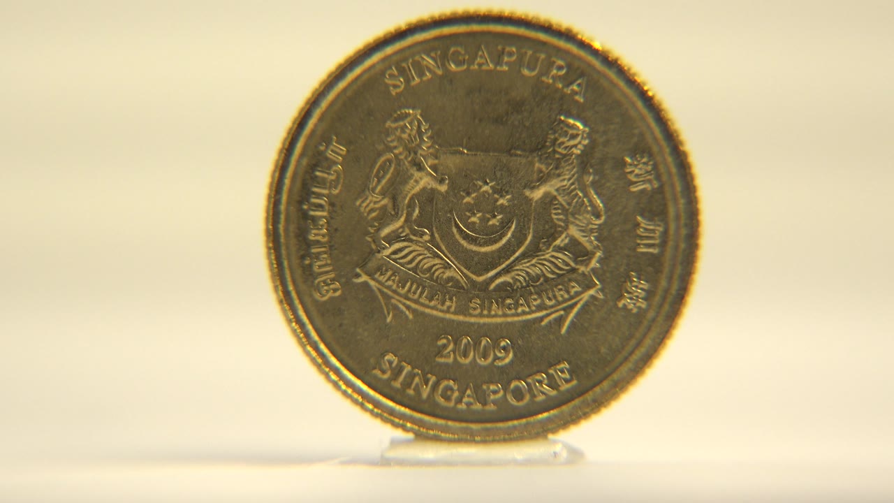 The coin remains nearly front-facing on the white surface, with raised lettering and metallic wear catching warm light, no visible people.