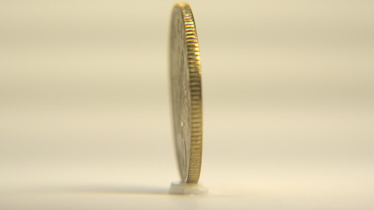 At 0:00, a Singapore coin is seen mostly from its thin edge while standing upright on a pale surface, with warm soft light and no visible people.