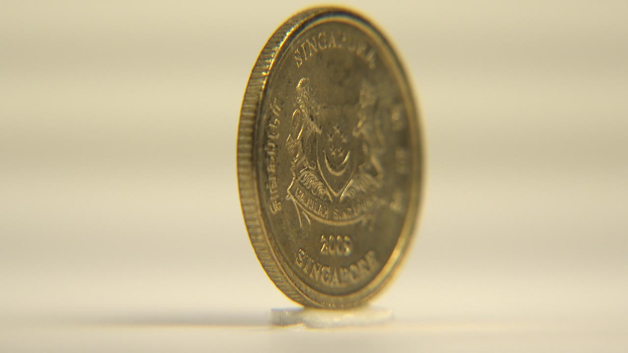 At 0:02, the coin face turns partly toward camera, showing Singapore text and crest detail in warm gold tones with no visible people.