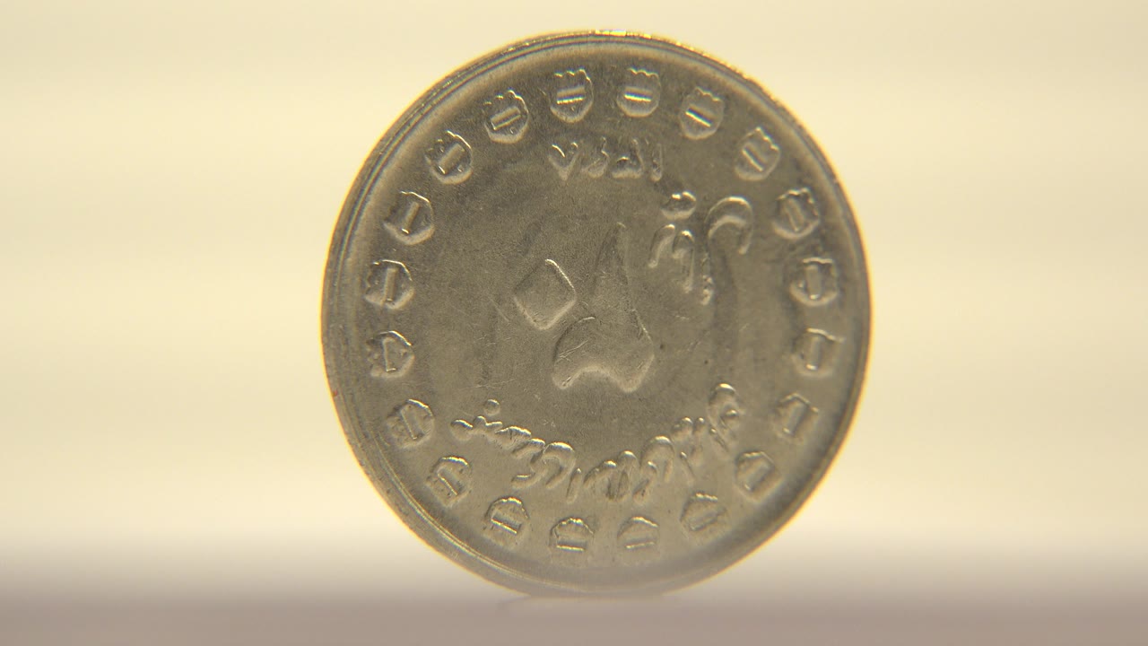 A worn metallic coin stands nearly face-on against a warm pale background, with raised script-like markings around the rim and no visible people.