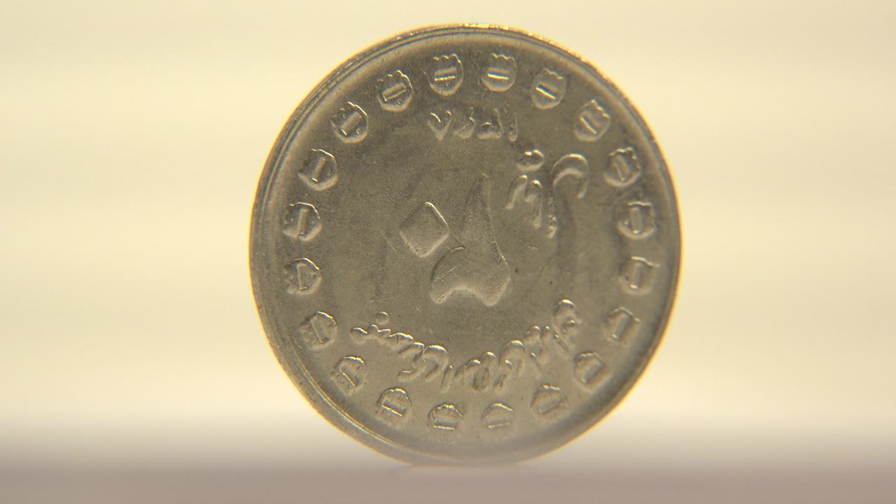 The coin is still mostly face-on, with soft warm light catching its worn raised symbols and the pale surface below, with no visible people.