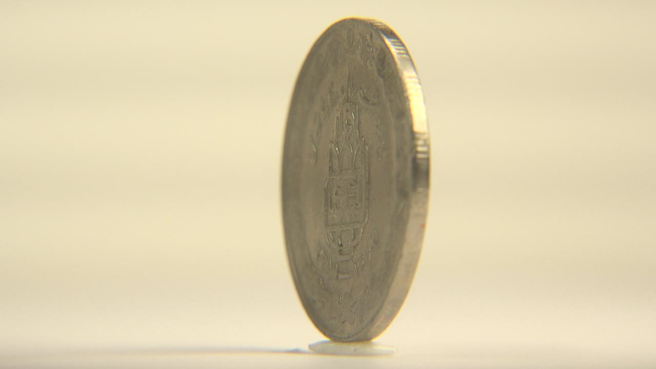 The coin remains upright in a narrow rim view, catching a bright highlight along one side against a soft cream background with no visible people.