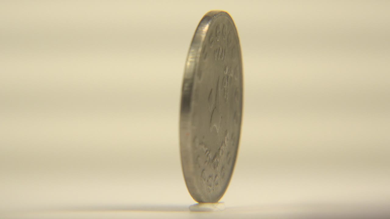 A slim side view of the upright coin shows the ridged edge and a small shadow at the base on a pale surface, with no visible people.