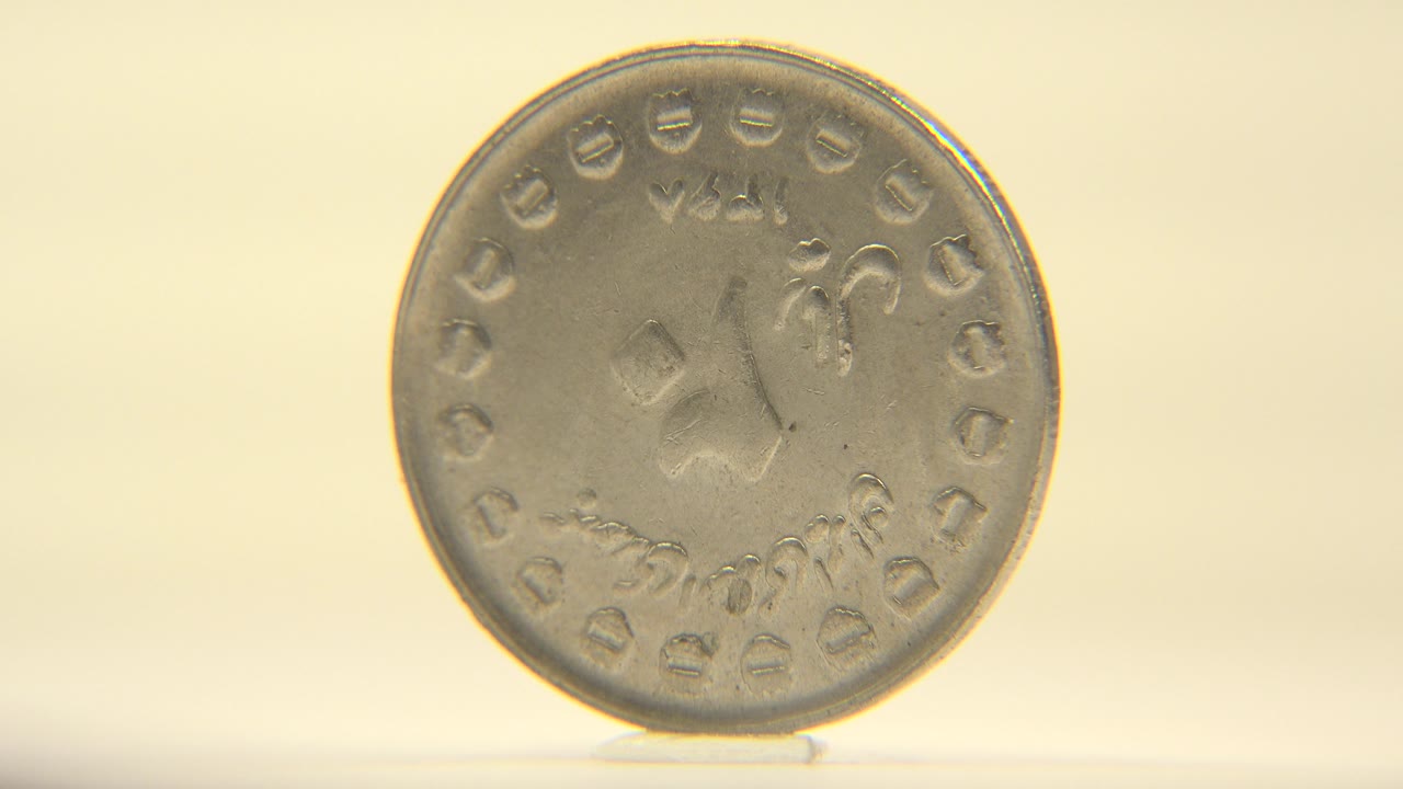 The coin is still upright with the front face visible and slightly shifted in angle, set against a plain cream backdrop with no visible people.