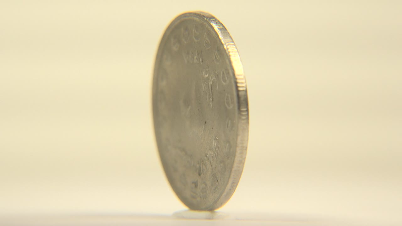 The coin turns toward a narrow edge view, showing its ridged side and blurred face detail on the cream surface, with no visible people.