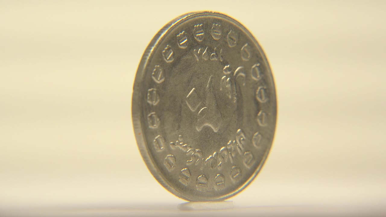 A silver-colored engraved coin stands upright near the left side of the frame on a pale surface, with warm cream background light and no visible people.