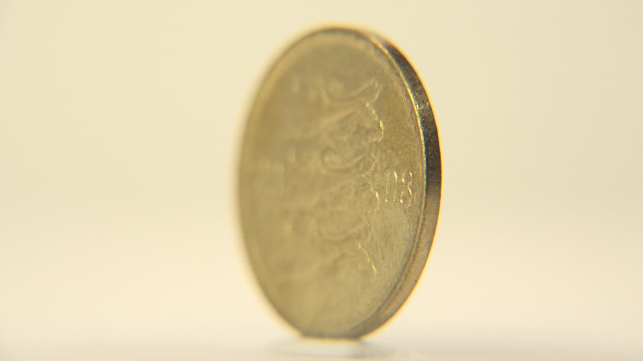 The coin has rotated to a slim edge view, showing the dark rim and thin profile against a warm blurred background with no visible people.