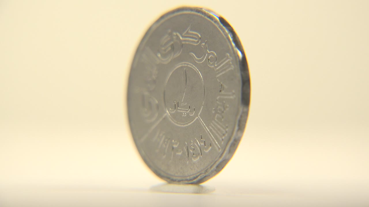 A silver-toned coin stands upright on a pale flat surface, showing raised script and a central numeral-like mark against a warm cream background with no visible people.