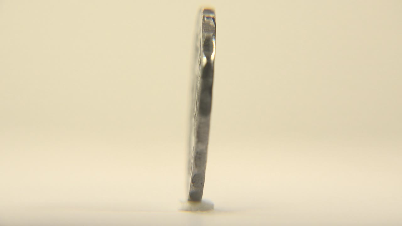 The rotating coin turns nearly edge-on, reducing to a narrow dark metal profile on the pale surface with a small contact shadow and no visible people.