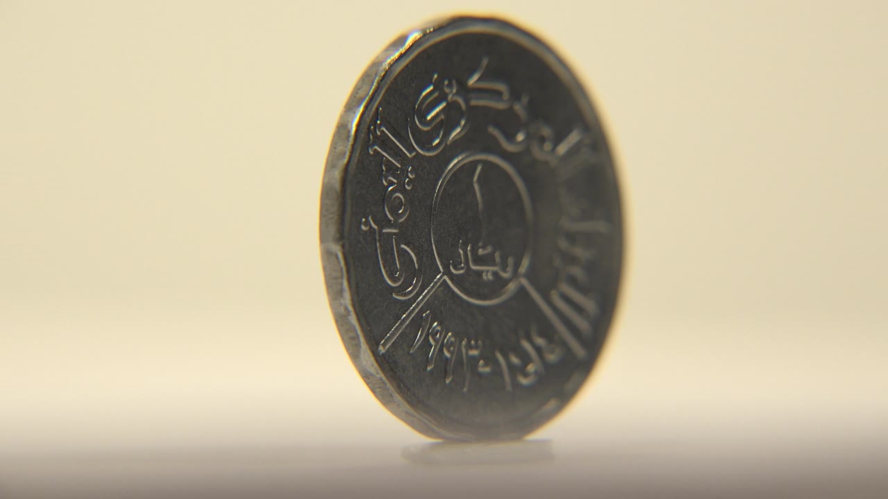 A dark metal coin stands upright at a slight angle on a pale flat surface, with Arabic lettering partly visible and no visible people.