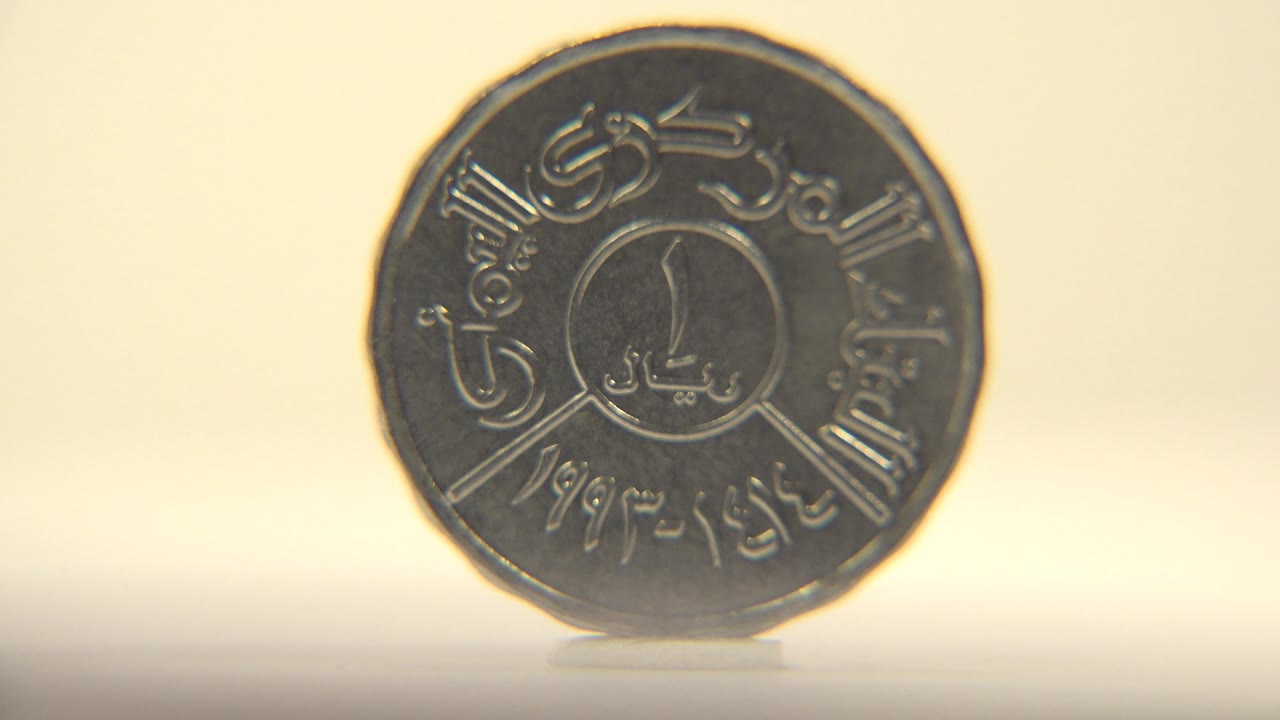The coin faces the camera more directly, with Arabic lettering around the rim and a small central design visible, and no visible people.