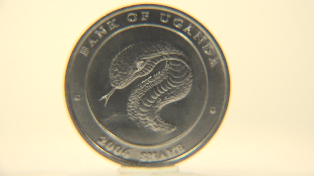 A silver Bank of Uganda coin faces the camera with a snake engraving visible on a warm cream background, no visible people.