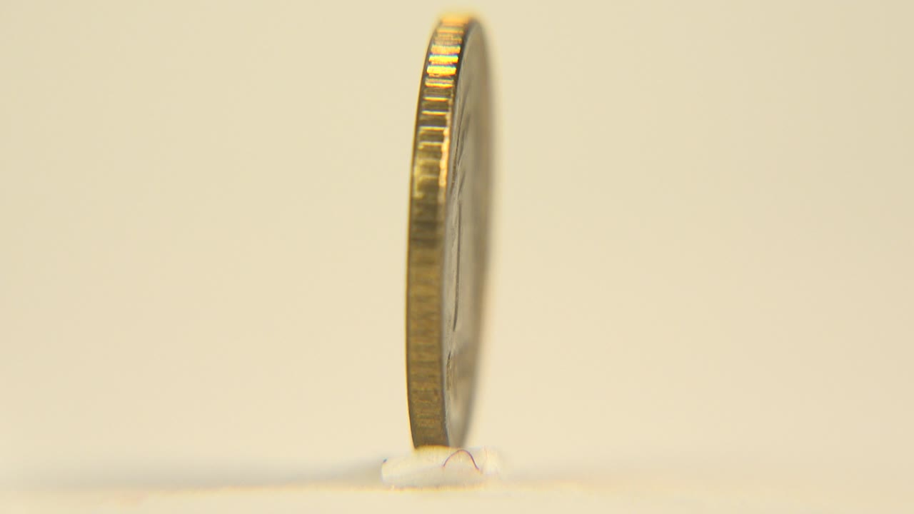 A gold-colored coin stands upright near the left side of the frame, shown mostly edge-on with a ridged rim, warm cream background, soft shadow, and no visible people.