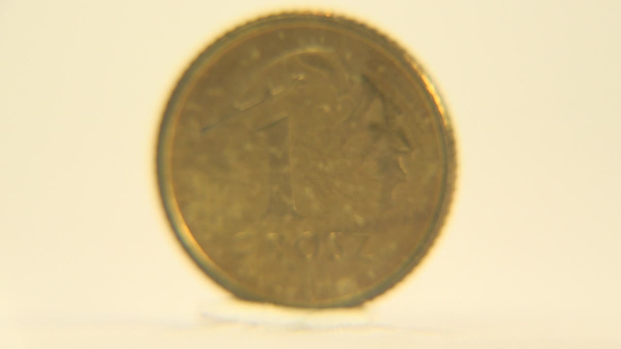 A gold-colored coin stands upright facing the camera on a pale reflective surface, with a warm cream background and no visible people.