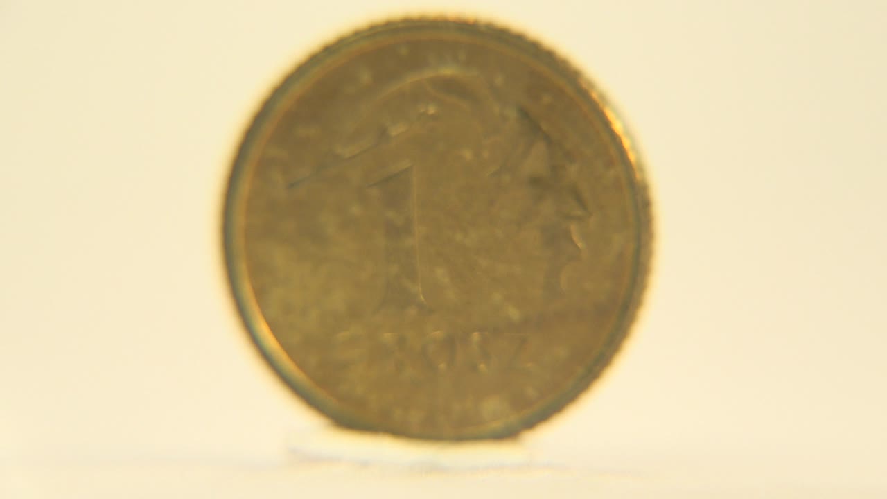 The upright gold-colored coin remains mostly front-facing, slightly darker in the center, on the same pale surface with no visible people.