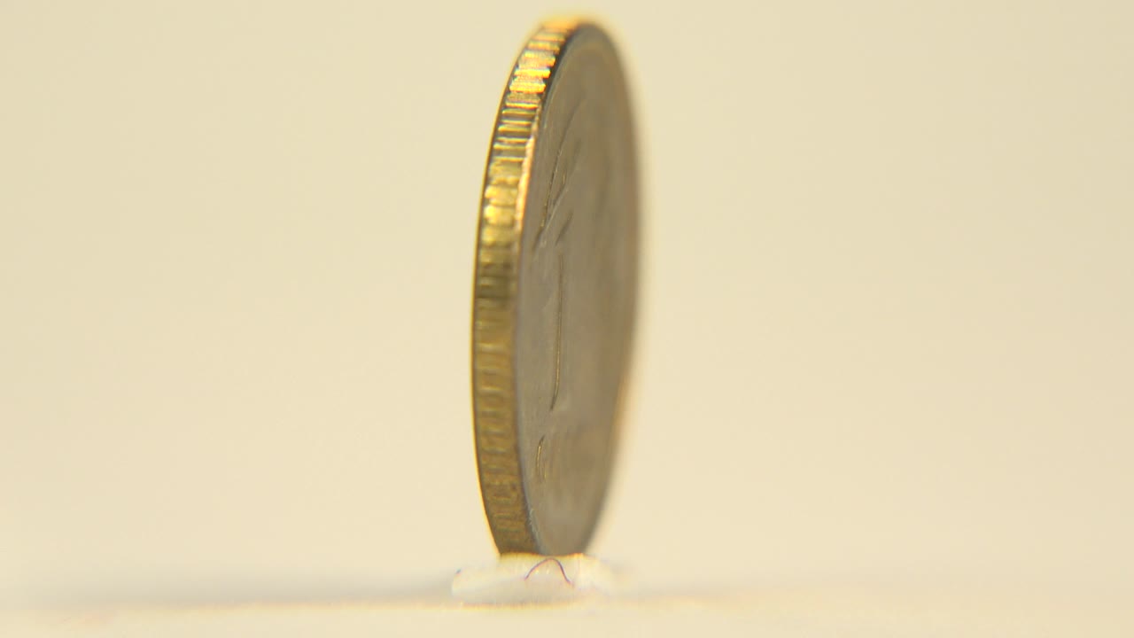 The coin turns almost edge-on, showing a narrow dark face and bright ridged rim while standing on the pale surface, with no visible people.