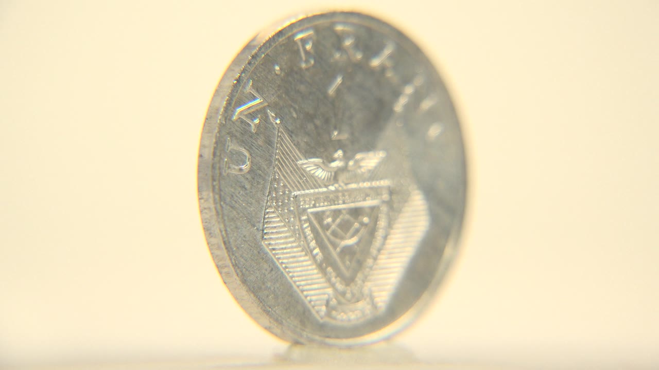 The coin faces the camera more directly in a soft close-up, with reflective metal, raised lettering, and a warm cream background with no visible people.