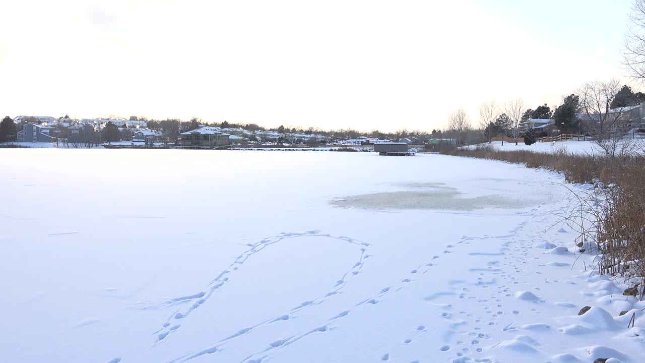 Snowy frozen lake surface in Hidden Lake, Colorado with a curved track crossing the foreground, a muted gray thawing ice patch beyond it, distant houses, and no visible people.