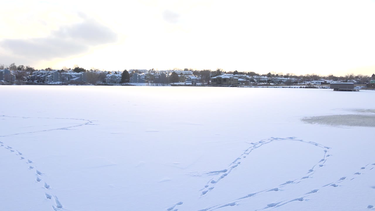 Early frame over Hidden Lake, Colorado, showing curved track marks crossing the white lake surface, a dark far tree line, small distant buildings, and no visible people.
