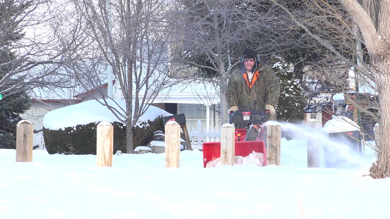 At 0:01, the man remains centered behind the red snow blower while a wide plume of snow blows to the right in front of the Hidden Lake residential background.