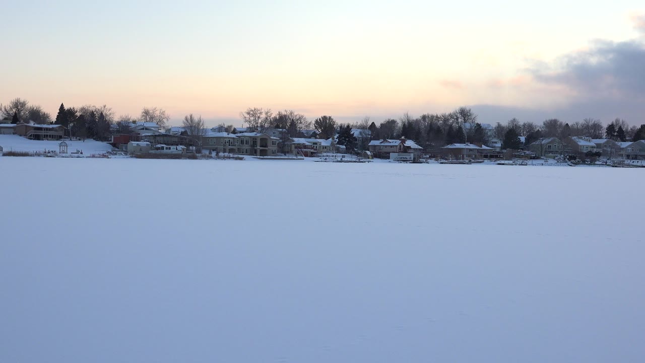 Snow-covered open foreground with distant houses and trees under a pale sunset sky.