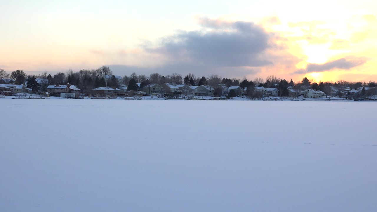 Wide view of a snow-covered lake or open frozen surface in Hidden Lake, Colorado, with distant houses, bare trees, a gray cloud, warm sunset light, and no visible people.