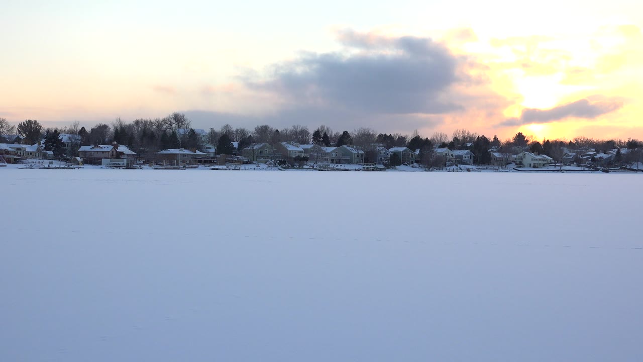 The second frame keeps the snowy lake foreground dominant, with Hidden Lake houses and trees along the horizon beneath a bright yellow sunset and no visible people.