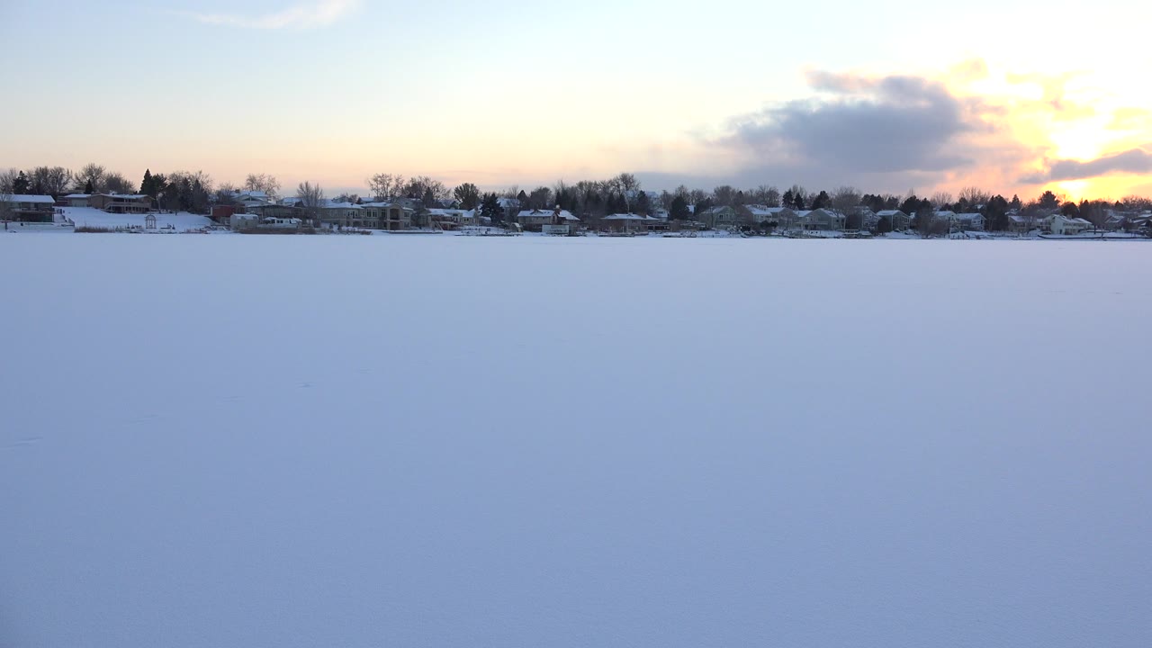 Wide snowy foreground with distant homes, trees, and a sunset sky.