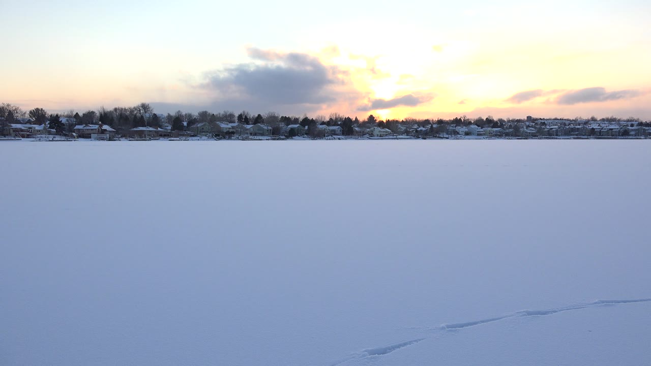 Snow-covered lake at sunset with distant shoreline and clouds.
