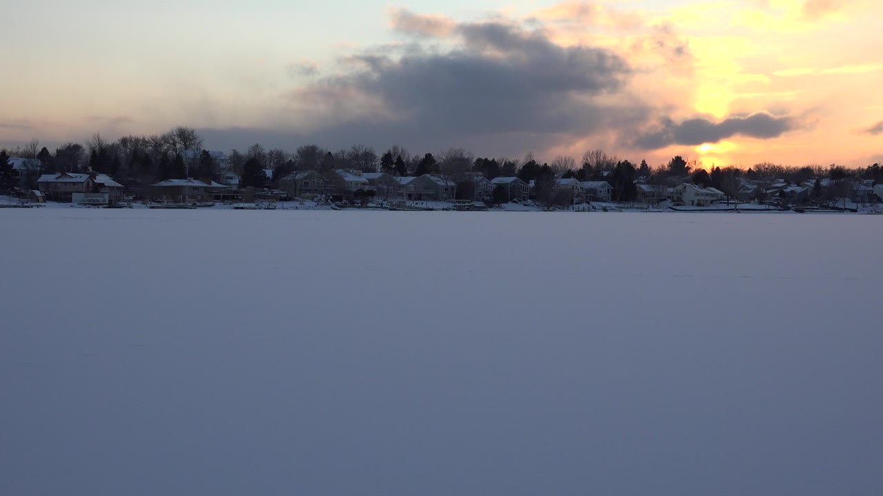 Wide view across Hidden Lake, Colorado with a blue snow-covered lake surface, distant houses and trees, orange sunset light behind clouds, and no visible people.