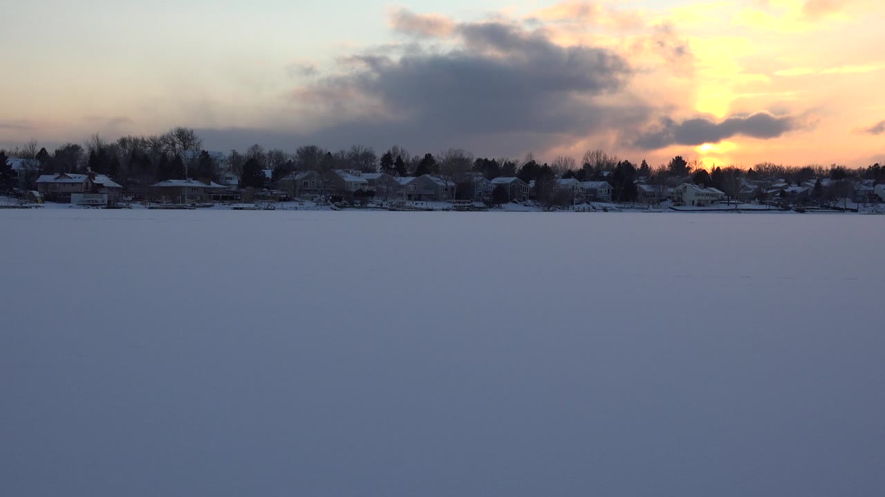 Hidden Lake, Colorado remains framed with the snowy lake filling most of the image, a narrow strip of houses and trees, and the sun glowing through darker clouds, with no visible people.