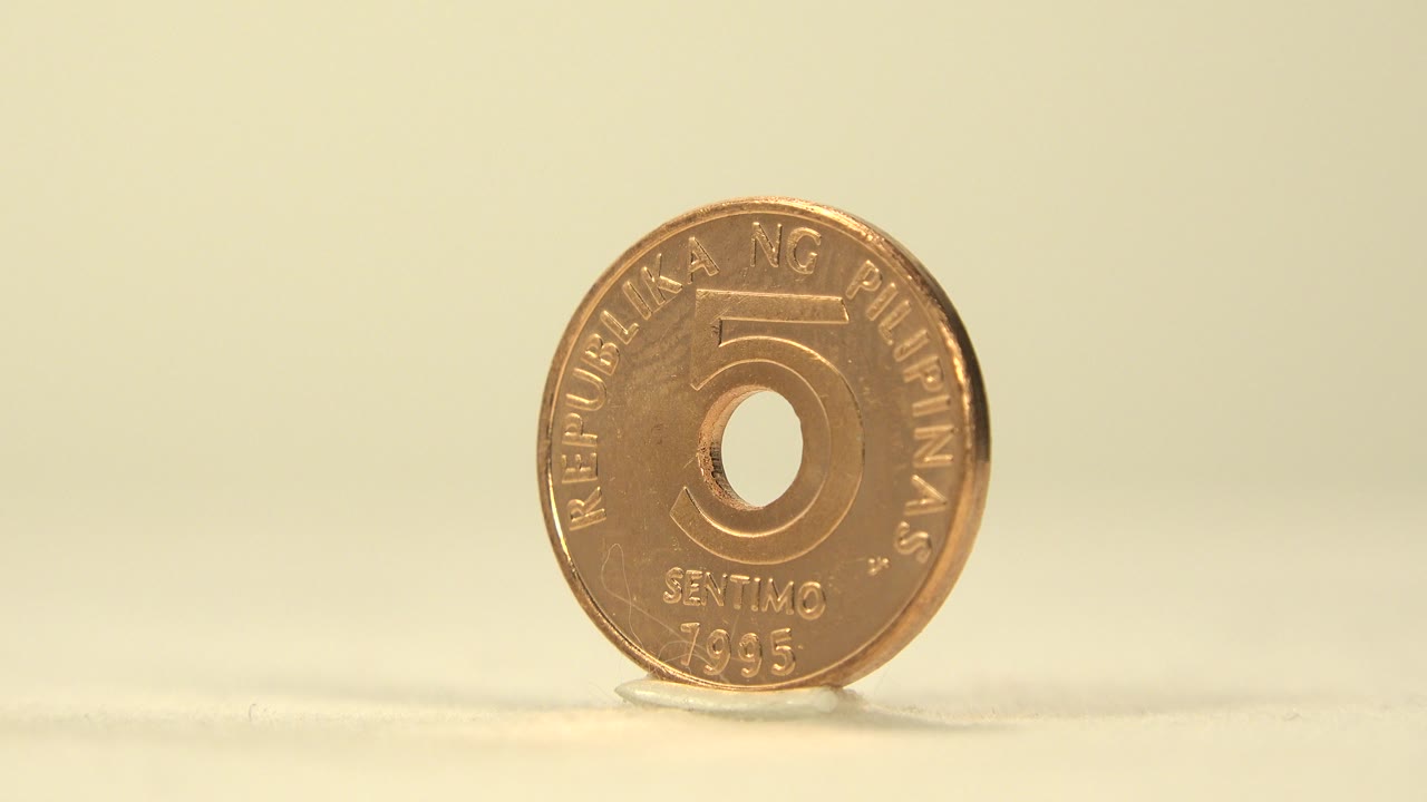 A copper-gold five centavo coin stands upright on a pale tabletop against a warm yellow background, no visible people.