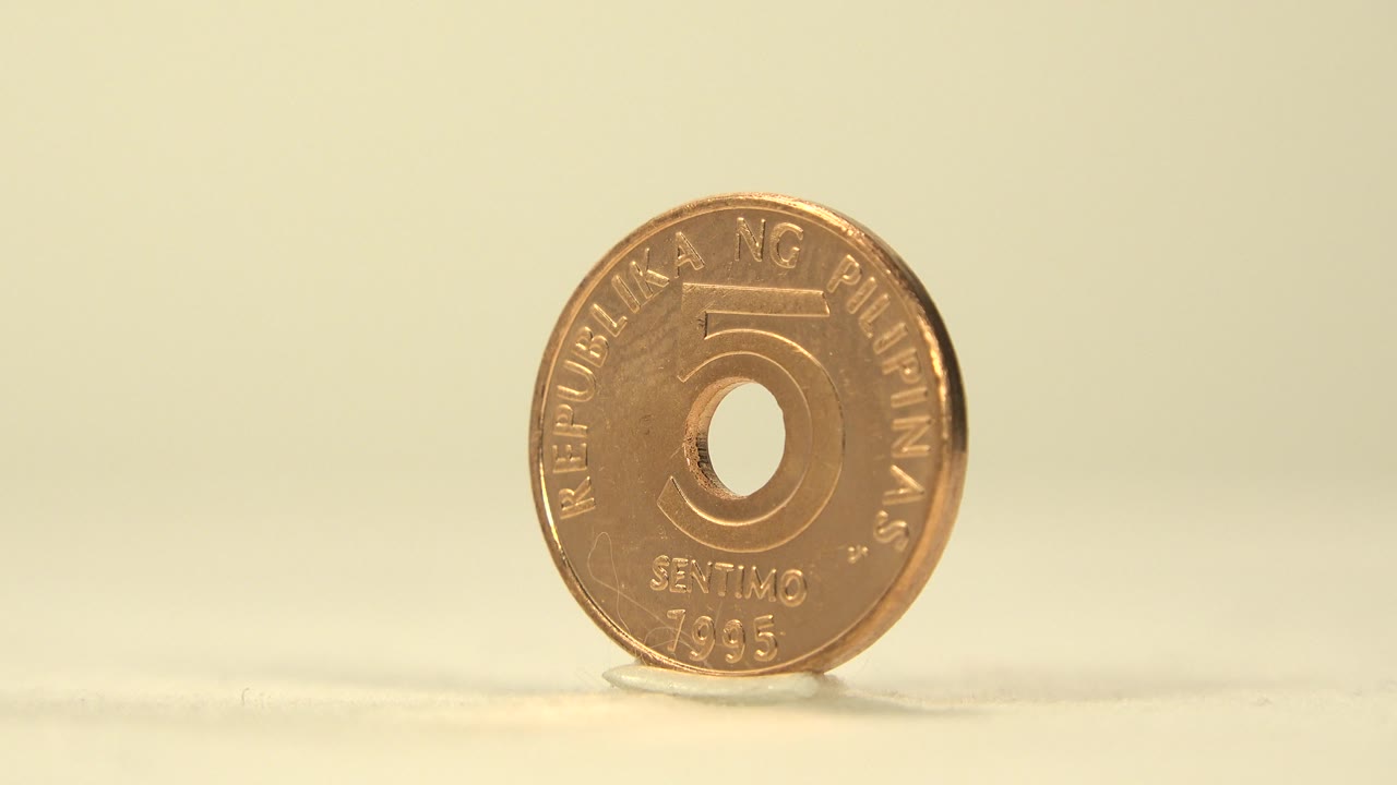 The coin remains upright and front-facing near the center of the frame, with the number 5 and circular cutout visible against the soft yellow background, no visible people.