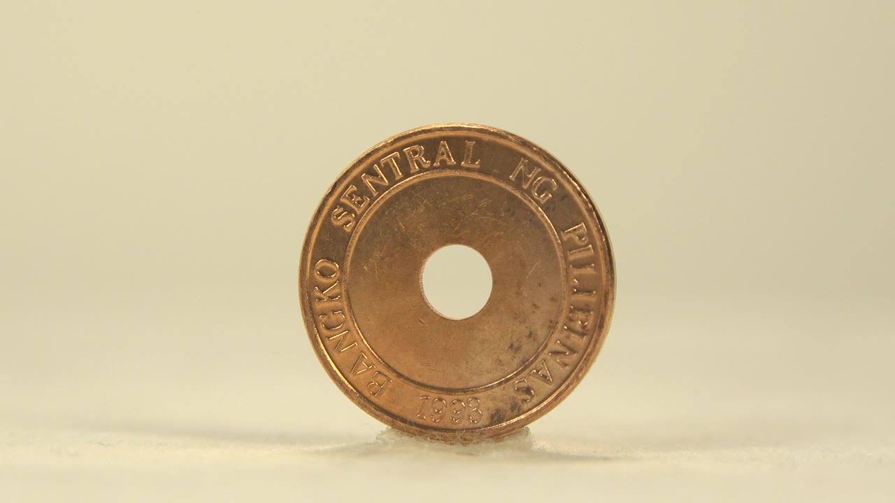 A holed 5 sentimo coin stands nearly face-on at the start of the clip, resting on one edge against a soft yellow-white background with no visible people.