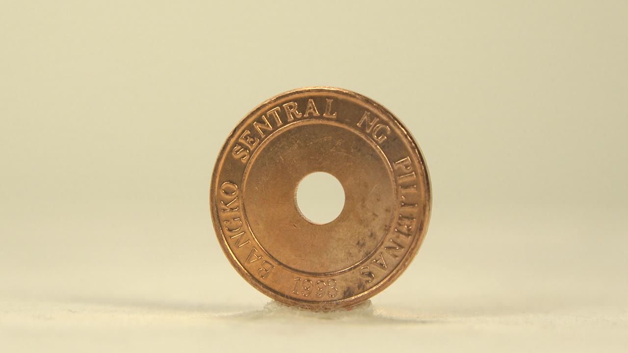 The coin remains upright and close to face-on, showing the central hole, raised rim lettering, warm copper-gold reflections, and no visible people.