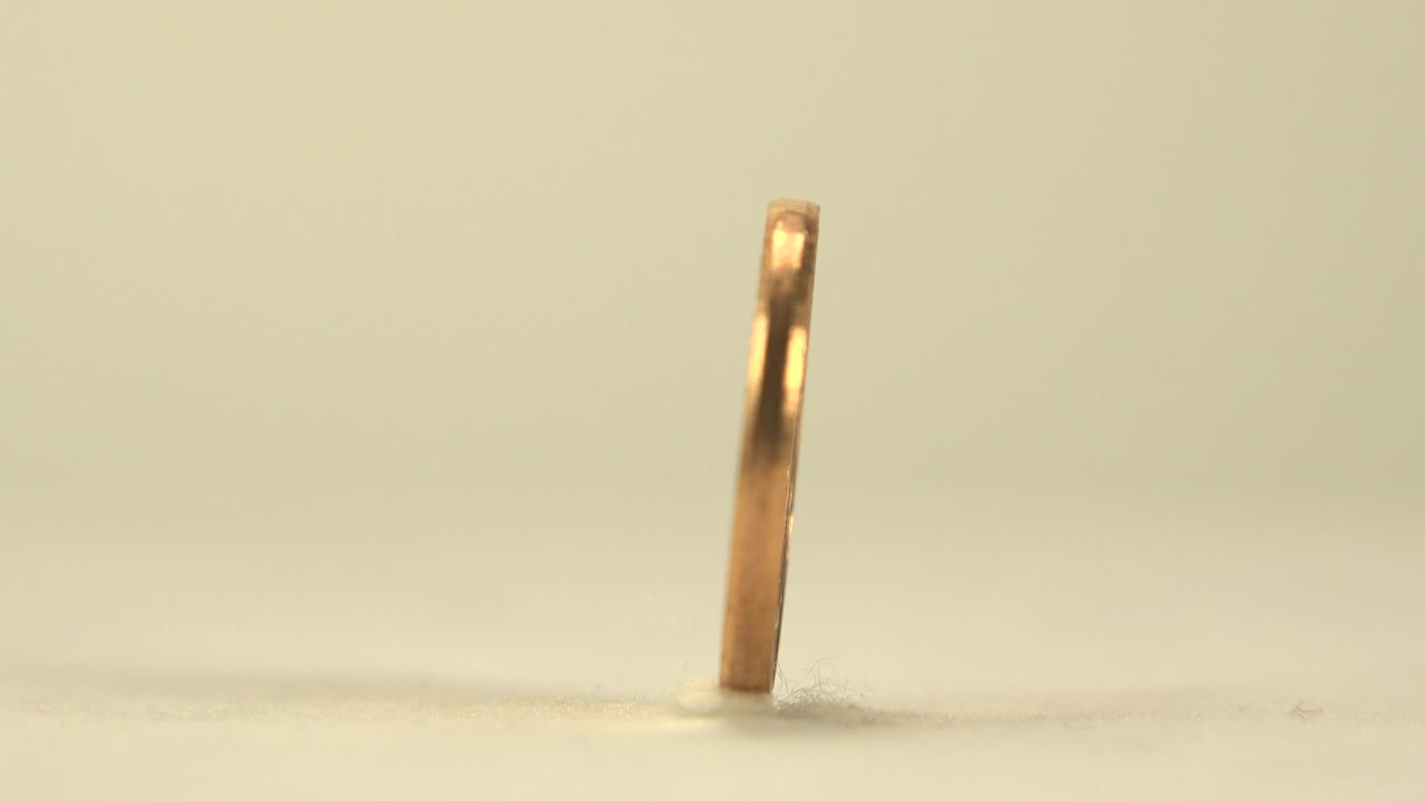 A golden coin with a center hole is seen almost edge-on while standing upright on a pale tabletop, with soft warm light and no visible people.