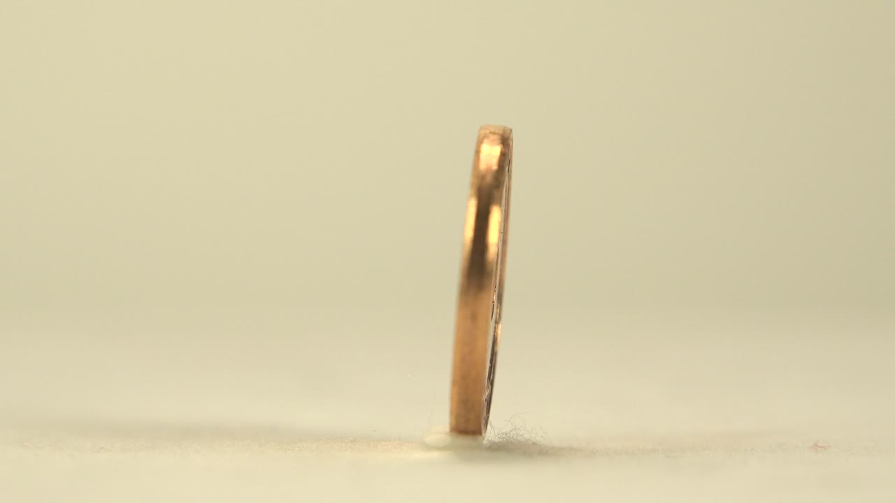 The coin remains upright in a thin edge view, catching a bright highlight along one side against the blurred pale background, with no visible people.