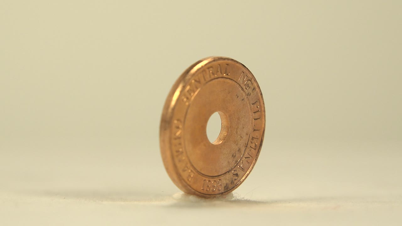 The coin has rotated to show more of its round face and central hole, with embossed lettering visible but not clearly readable throughout, and no visible people.