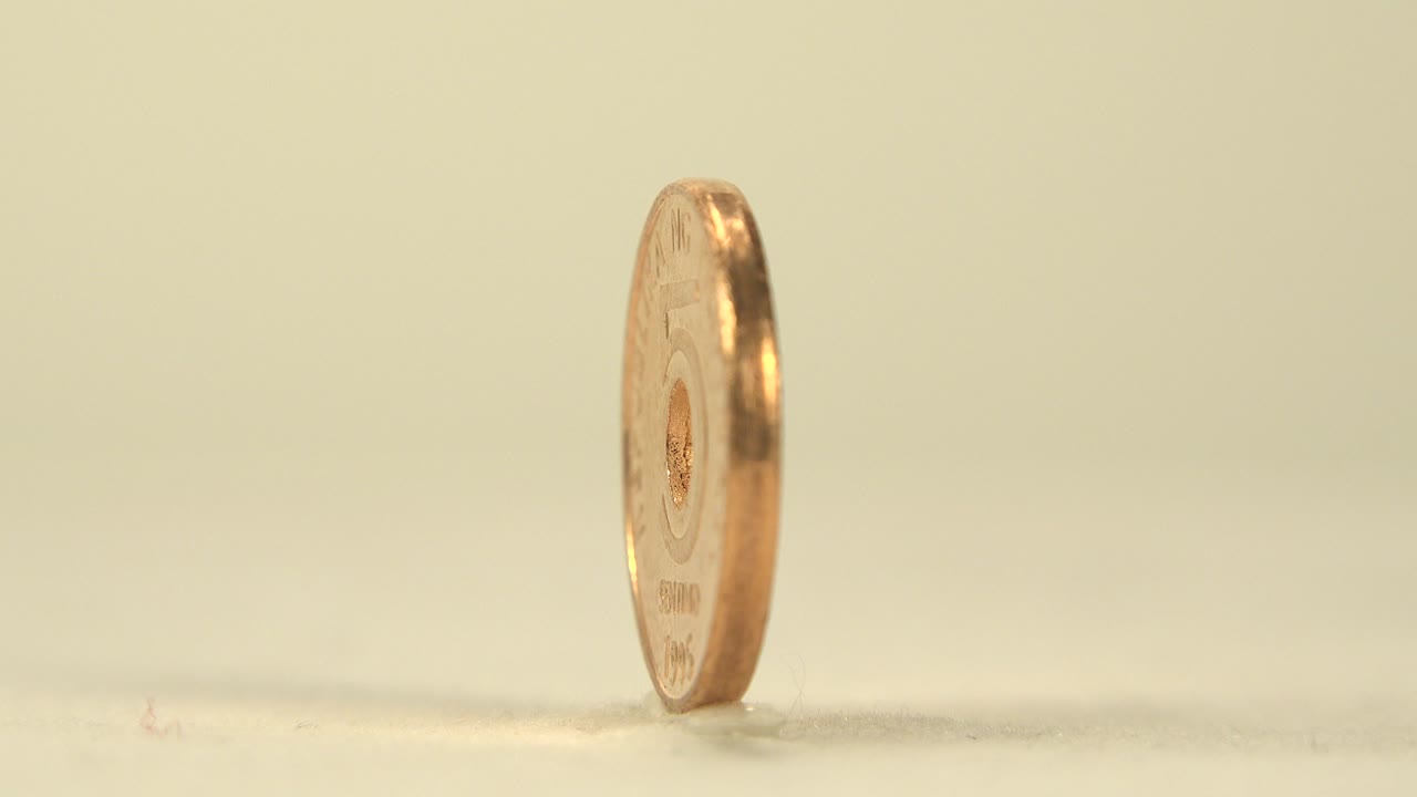 A copper-gold coin with a drilled center stands nearly edge-on on a pale tabletop against a soft cream background, with no visible people.