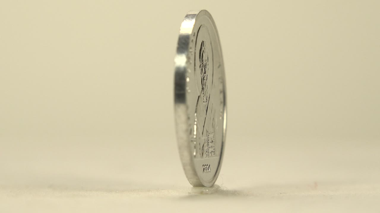 The coin remains upright and slightly angled, showing a reflective rim and faint face detail on a pale surface with no visible people.